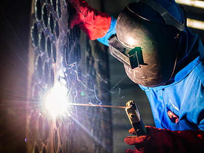 Ein Schweißer trägt eine Schweißmaske und schweißt an einem Metallstück.